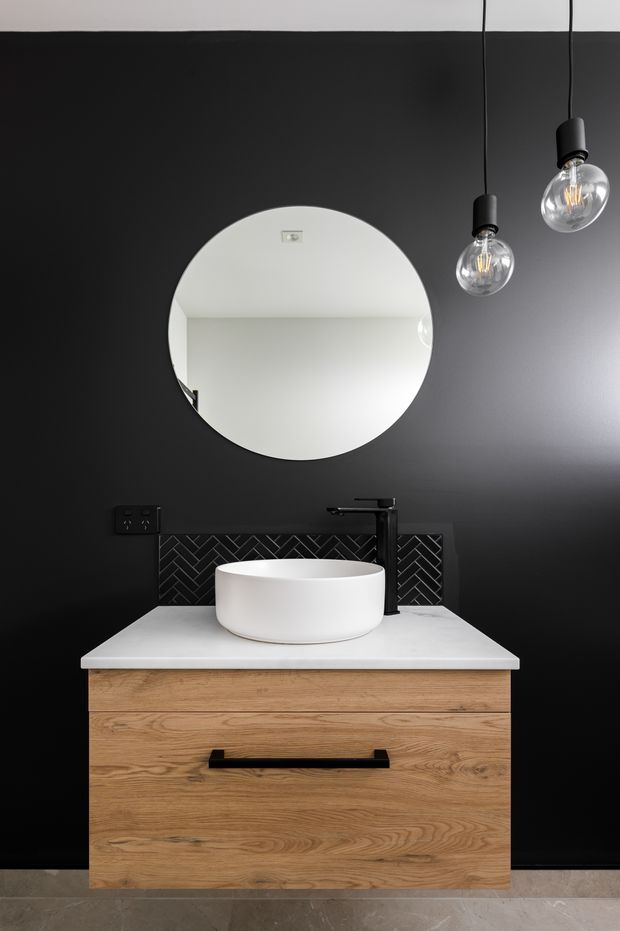 A contemporary bathroom vanity composition featuring a wall-mounted natural wood drawer cabinet, white vessel basin, black tapware, white benchtop, and dark herringbone tile splashback against a matte black wall. A large round mirror and two hanging globe pendant lights provide contrast and illumination. Grey stone-look floor tiles are visible at the base.