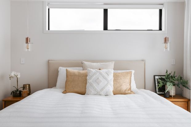 A bright, contemporary bedroom viewed straight on, featuring a neatly made bed with white bedding, beige and textured decorative cushions, a low upholstered headboard, timber bedside tables, wall-mounted pendant lights, and a wide horizontal black-framed window. Neutral finishes and greenery create a calm, refined atmosphere.