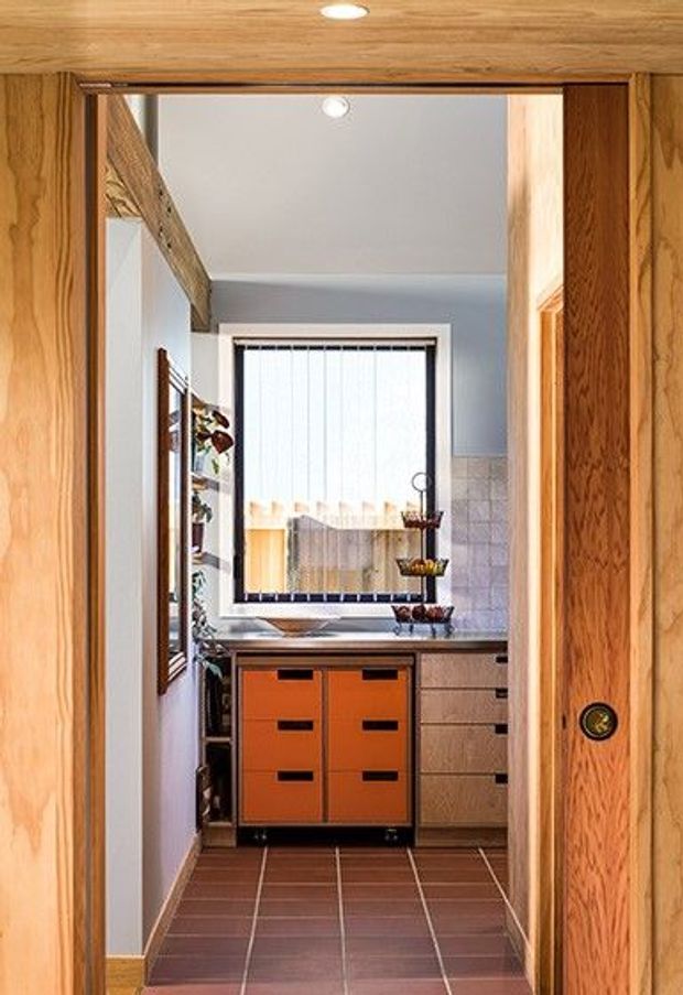 A compact kitchen or utility workspace viewed through a timber-framed doorway. The room features warm wood wall and ceiling finishes, a dark tiled floor, pale walls, a tiled backsplash, a window with vertical blinds, and a distinctive orange built-in drawer unit beneath the benchtop. Open shelving and timber cabinetry are visible along the sides.