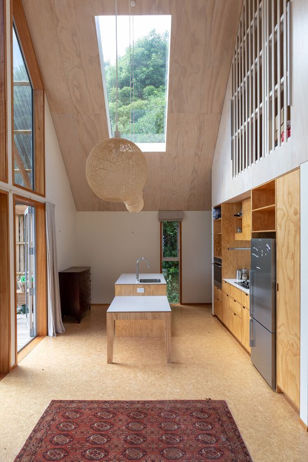 A bright, double-height kitchen viewed from an open living area, featuring extensive warm plywood cabinetry, a central island with an integrated sink, cork-toned flooring, tall glazing, and a large roof skylight. A timber slatted balustrade overlooks the kitchen from an upper level, while a woven pendant light and patterned rug add warmth and texture.