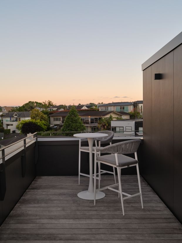 A compact elevated outdoor terrace or balcony overlooking a suburban neighbourhood at sunset. The space features dark privacy walls and cladding, grey weathered timber decking, and a small round high table with two woven bar stools. The terrace has a contemporary, minimalist appearance with muted neutral tones and open suburban views.