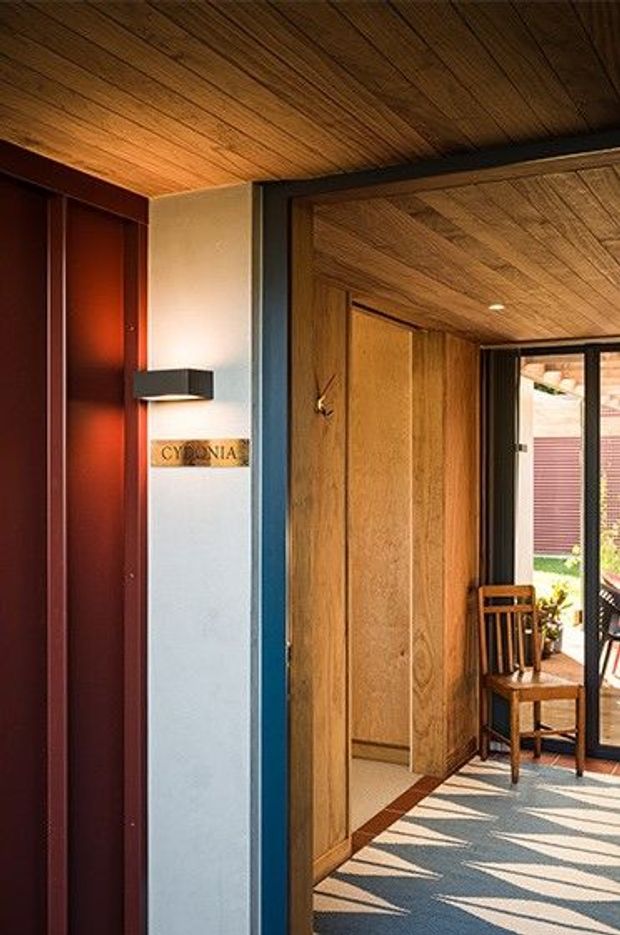 A warmly lit interior entryway or hallway featuring extensive timber lining on the ceiling and walls, custom timber panelled doors, and dark red vertical wall panelling. A narrow white display or built-in element carries a small sign, while a wooden chair sits beside full-height glazing opening toward an outdoor area. Strong sunlight creates geometric shadows across the floor, with dark blue-black trim providing contrast.