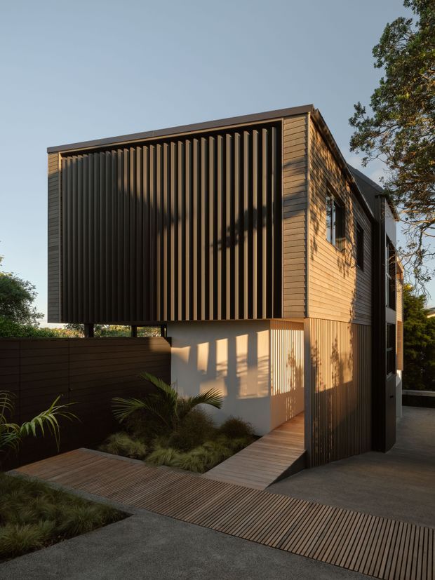 Exterior view of a narrow, multi-level contemporary home with a strong rectilinear form. The facade combines horizontal timber cladding with dark vertical screening and a projecting upper volume, creating privacy and patterned shadows. A timber boardwalk crosses a landscaped courtyard and leads toward the entry, with a concrete driveway and tall boundary fencing in the foreground.