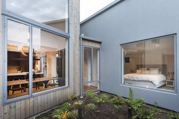 A contemporary residential courtyard viewed from outside, with a small planted garden between two wings of the home. Floor-to-ceiling glazing reveals a warm dining and kitchen area on the left, while a large bedroom window on the right shows a neatly made bed and bedside tables. The facade combines pale vertical timber cladding with smooth grey rendered walls and slim dark window frames under a simple pitched roofline.