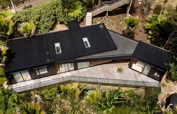Aerial view of a contemporary single-storey dwelling nestled in dense landscaped vegetation. The building has a dark metal pitched roof with two skylights, extensive glazing, and a broad timber deck wrapping around the front and side. A raised timber walkway and stairs connect through the garden behind the house.