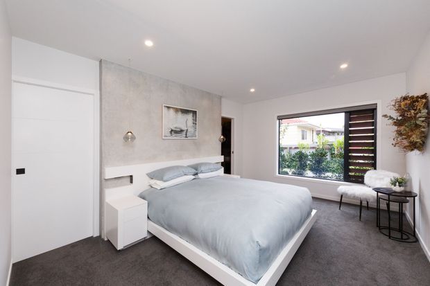 Bright contemporary bedroom with a low white platform bed dressed in pale grey bedding, dark grey carpet, white walls, and a textured grey feature wall behind the bed. The room includes integrated bedside storage, recessed ceiling lights, a pendant, a framed artwork, a large window with dark timber screening, and a small seating area with side tables and dried wall foliage.