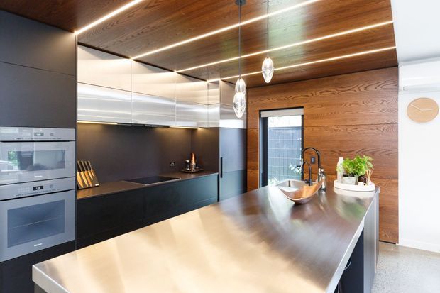 A contemporary kitchen with dark cabinetry, integrated appliances, a large stainless-steel island and timber-lined ceiling and feature wall. Linear recessed lighting and pendant lights illuminate the open, streamlined space, with a sink and tap positioned at the island.
