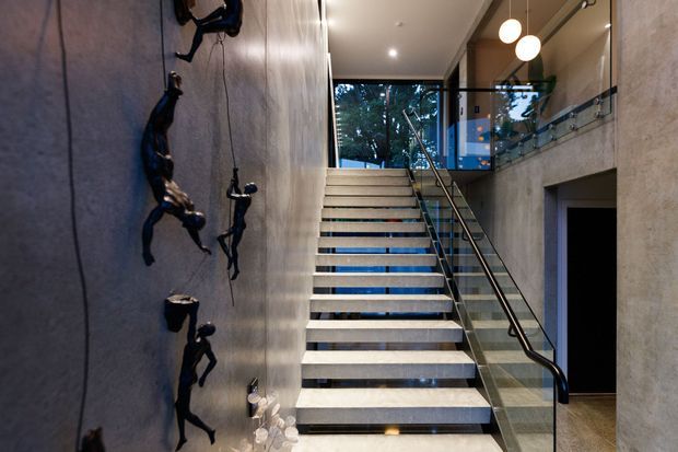 A dramatic modern stairway rises through a narrow double-height interior. Floating pale stone or concrete treads contrast with dark riser gaps and a glass balustrade with black metal handrail. The left wall has a textured dark finish with sculptural climbing figures, while recessed ceiling lights and spherical pendant lights illuminate the space. Glazing at the upper landing reveals trees outside.