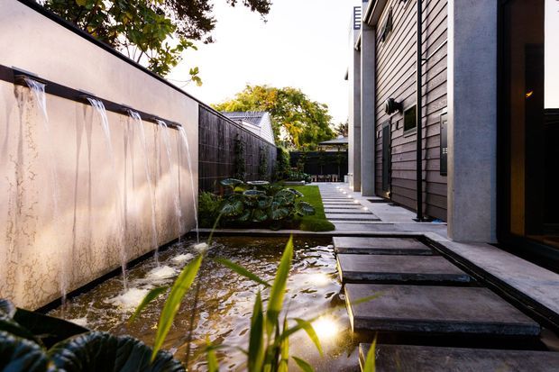 A narrow landscaped courtyard beside a contemporary building, featuring a shallow reflective water feature with wall-mounted cascades, stepping-stone paving, dark timber or composite wall cladding, and lush planting. The walkway leads toward a small garden enclosure, creating a tranquil outdoor passage.