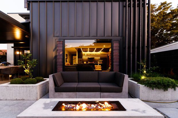 Contemporary outdoor entertaining area with a built-in linear fire pit set into a pale concrete table or hearth, facing a dark upholstered outdoor sofa. The space is framed by dark vertical metal cladding, raised concrete planters, architectural lighting, and a large opening revealing a warmly lit interior lounge or kitchen beyond.