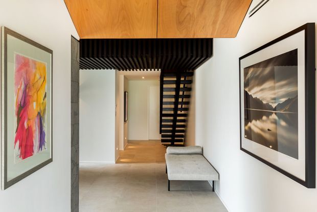A contemporary residential hallway or entry passage with a sculptural open-riser staircase, black structural detailing, warm timber ceiling panels, pale walls, and light flooring. Framed artwork and a streamlined grey upholstered bench add visual interest while preserving a clean, gallery-like feel.