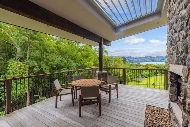 Covered timber deck and outdoor entertaining area with a round dining table and chairs, stone-clad wall, dark timber posts and railing, and a large roof skylight. The elevated deck overlooks lush bushland and a broad mountainous valley landscape.