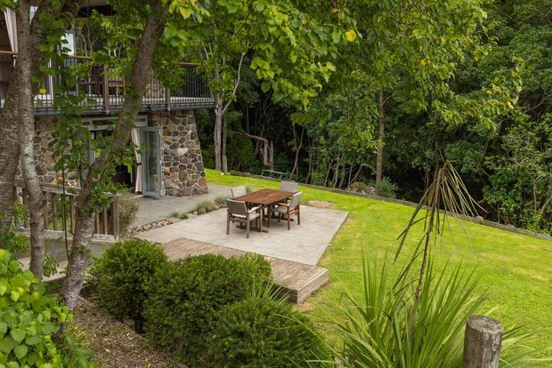 A landscaped outdoor terrace beside a stone-clad multi-level home, with a timber dining table and chairs on a pale paved patio, surrounded by dense mature greenery and lawn. Timber decking steps connect the terrace to the garden, and an upper balcony is visible through the trees.