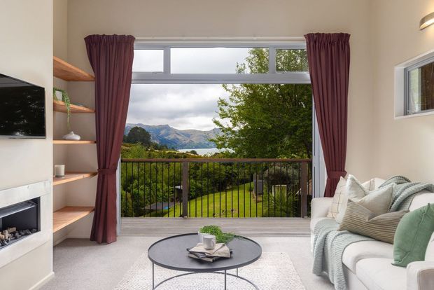 Bright contemporary living room with a large window and glass door opening to a balcony overlooking a landscaped garden and mountainous landscape. The room features a pale upholstered sofa, round black coffee table, built-in timber shelving, a wall-mounted television, fireplace, light carpet, and burgundy curtains.