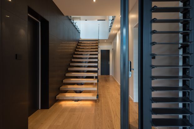 Interior view of a contemporary stair hall with floating timber treads, dark metal supports and handrails, and warm timber flooring. Dark built-in wall panelling and cabinetry line the left side, while a glazed and louvred dark-framed opening is visible on the right. Recessed lighting and a double-height stair void create a refined, modern atmosphere.