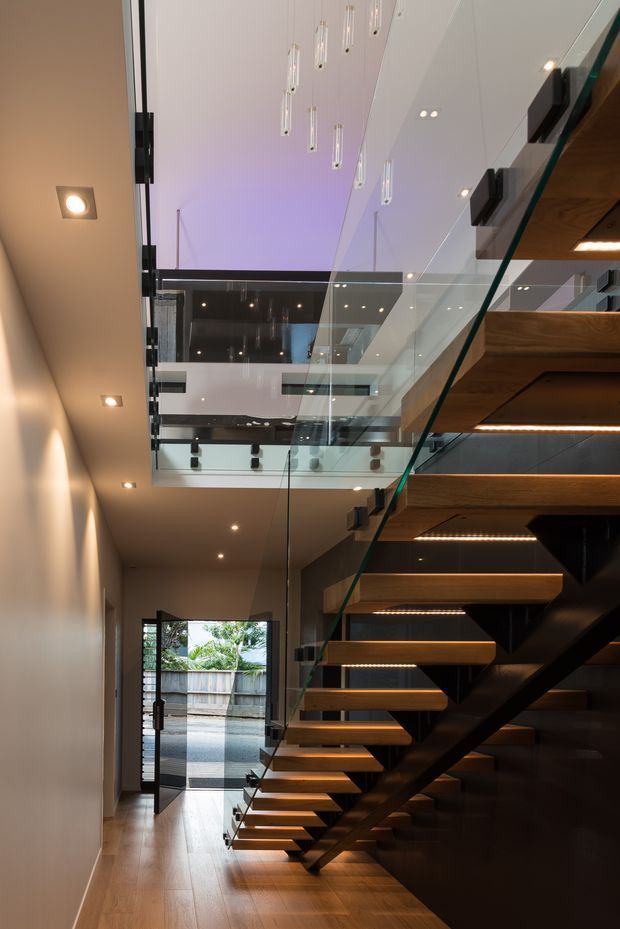 A tall contemporary interior hallway dominated by a floating timber staircase with dark structural supports, clear glass balustrades, and integrated LED lighting beneath each tread. The stairway rises through a multi-level void with glass-edged landings, recessed ceiling lights, and suspended cylindrical pendant lights. Warm timber flooring and stair treads contrast with white walls and dark joinery, while an open doorway leads outside at the end of the hall.