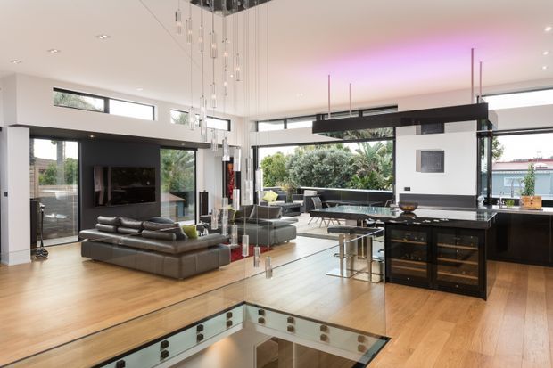 Open-plan contemporary living, dining and kitchen space with expansive glazing, warm timber flooring, a dark kitchen island and bar cabinetry, a sunken stair opening in the foreground, and a dramatic cluster of suspended pendant lights. The room has a bright, airy feel with subtle purple cove lighting and views to greenery outside.