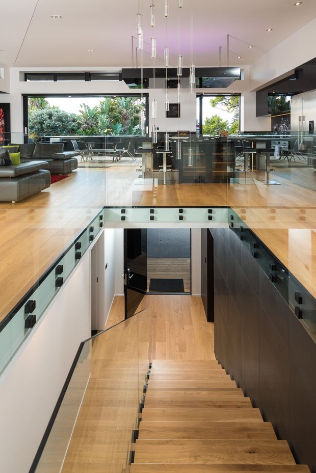 A contemporary open-plan living area viewed from an upper-level landing, with a dramatic stairwell descending through the centre. The space features warm timber flooring and stairs, extensive black-framed glazing, glass balustrades, dark built-in cabinetry, grey sectional seating, and a cluster of slender pendant lights. Lush garden planting is visible through the windows, while a kitchen or bar area sits beyond the living zone.