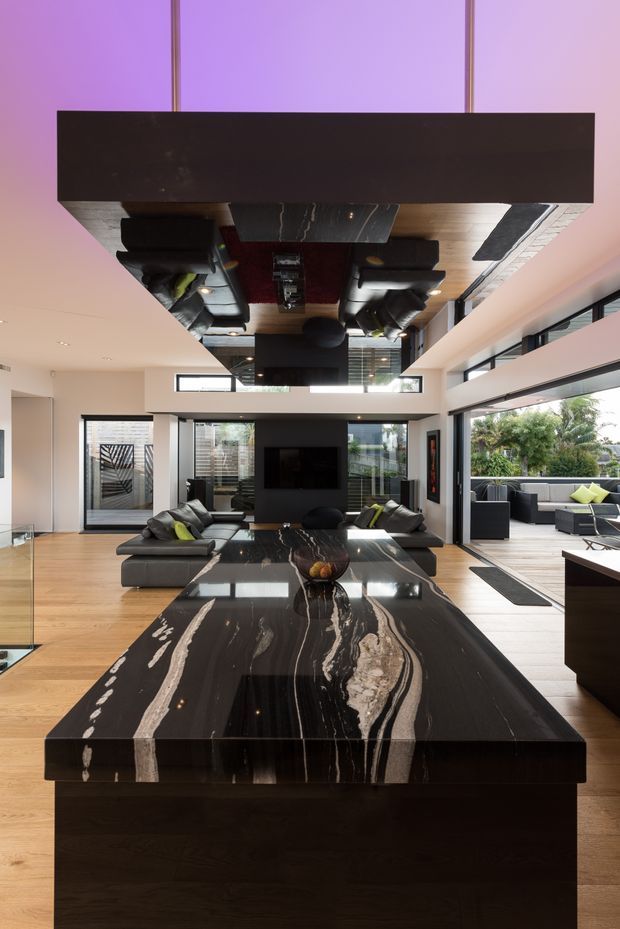 A dramatic contemporary open-plan living area viewed from a kitchen island. The foreground features a large black marble island benchtop with striking white veining and dark timber cabinetry. Beyond it is a sophisticated lounge with black sofas, a central television wall, extensive glazing, timber flooring, and a mirrored ceiling detail reflecting the room and pendant lighting. The space opens toward an outdoor seating area and garden.