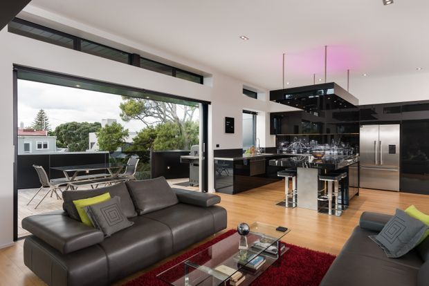 Open-plan contemporary living, dining and kitchen interior with polished dark cabinetry, a large black island, stainless-steel appliances, timber flooring and expansive sliding glass doors opening to a terrace. Dark leather sofas and a red rug define the living area, while a black-framed clerestory and recessed ceiling lighting add architectural detail. The space has a strong indoor-outdoor connection and overlooks neighbouring greenery and rooftops.