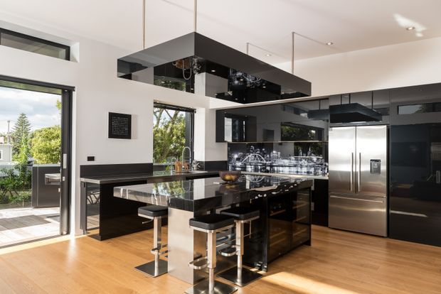 A contemporary open-plan kitchen with a glossy black island, integrated black cabinetry, reflective overhead rangehood, stainless-steel appliances, and bar seating. Warm timber flooring contrasts with white walls and large glazed openings that bring in abundant daylight and views of greenery.