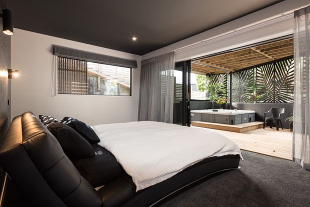A contemporary bedroom with a large black upholstered bed, white bedding, dark carpet, charcoal ceiling, and sheer floor-to-ceiling curtains. Sliding glass doors open directly onto a timber-decked outdoor terrace featuring a spa, outdoor seating, timber overhead screening, and patterned privacy panels.