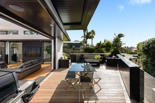 Contemporary outdoor entertaining terrace with a timber deck, outdoor dining table and chairs, built-in dark cabinetry or counter, lounge seating, and a barbecue. Large sliding glass doors connect the deck to an open-plan interior living area. The space overlooks a sunny suburban landscape with palm trees and neighboring homes.