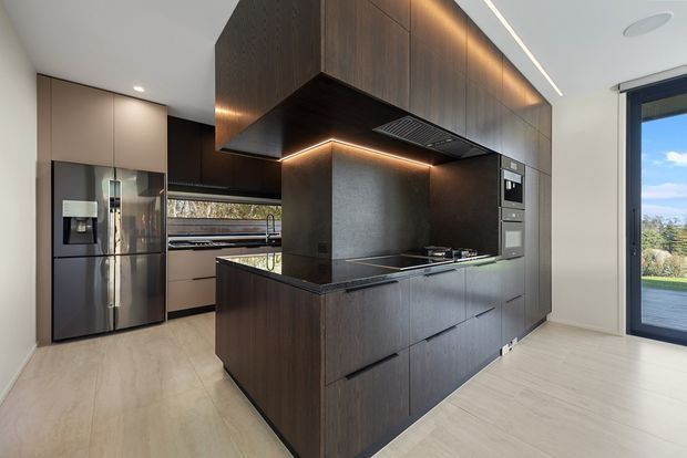 Contemporary kitchen featuring a large dark timber island with integrated cooktop, dark stone benchtops, full-height timber cabinetry, built-in appliances, warm under-cabinet lighting, and a stainless-steel refrigerator. Pale wood-look flooring and a large glazed door create a bright, refined interior.