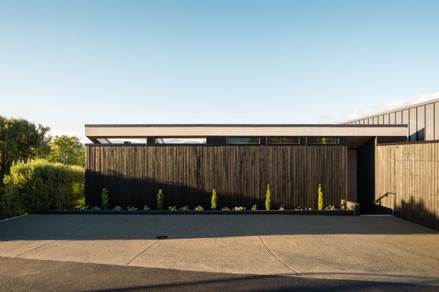 A low, contemporary residential facade viewed from the driveway. The building features dark vertical timber cladding, a long horizontal roof edge with clerestory-like glazing beneath it, and a contrasting pale timber-clad side volume. A broad concrete-paved forecourt and simple landscaped planting occupy the foreground.