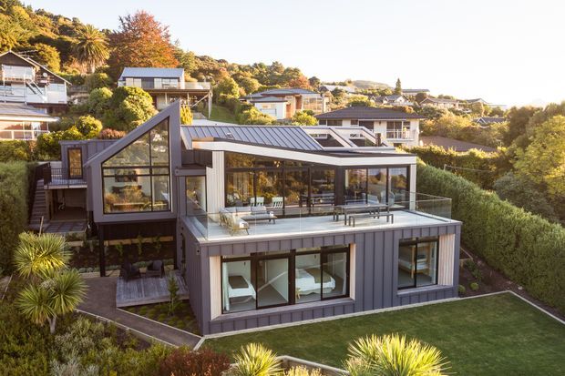 Aerial exterior view of a contemporary multi-level hillside residence with dark vertical cladding, extensive glazing, a prominent sloped roof form, and a large upper-level glazed living area opening onto a glass-balustraded terrace. Landscaped gardens and neighbouring hillside homes surround the property.