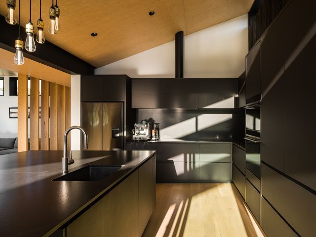 A sophisticated contemporary kitchen with matte dark cabinetry, dark stone benchtops, integrated appliances, and a large island with a curved chrome tap and undermount sink. Warm timber ceiling panels and flooring contrast with the black joinery, while exposed pendant bulbs and vertical timber screening add architectural character. Sunlight streams across the cabinetry and floor, and a living area is partially visible beyond the kitchen.