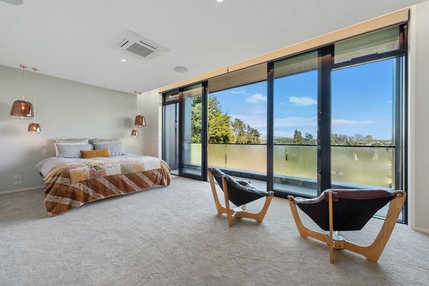 Bright contemporary bedroom with a low-profile bed, neutral carpet, copper pendant bedside lights, and two sculptural timber-framed lounge chairs. Full-height black-framed glazing opens onto a balcony with frosted lower panels and views of trees and blue sky.