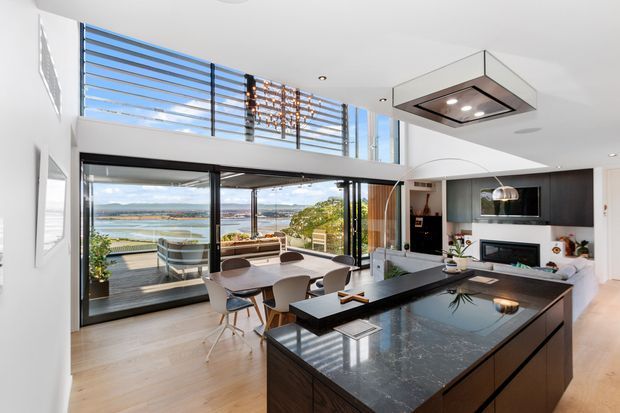 Bright contemporary open-plan kitchen and living area with a large dark stone island, integrated cabinetry, dining table, and expansive glazed doors opening to a balcony. The room has a double-height feel with upper clerestory glazing and views toward a waterfront landscape.