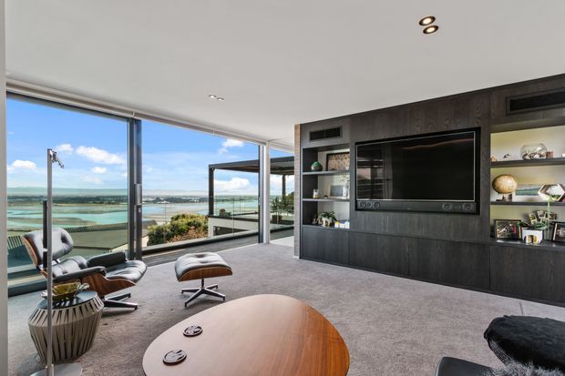 Contemporary living room with expansive floor-to-ceiling glazing overlooking a coastal waterfront landscape. The room features a dark timber entertainment wall with built-in television and open shelving, a grey carpeted floor, a sculptural lounge chair with ottoman, and a rounded timber coffee table. A covered outdoor terrace is visible beyond the glazing.