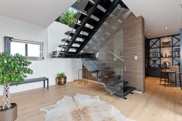 A bright contemporary interior featuring a dramatic open-riser staircase with black steel stringers, dark treads, glass balustrades, and a timber-clad feature wall. The staircase connects an open entry or living area to an upper level, with light timber flooring, white walls, a sculptural potted plant, bench seating, and a visible home bar or display joinery area in the background.