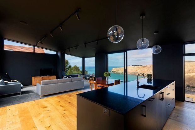 A dramatic open-plan kitchen and living area with dark walls and ceiling, extensive floor-to-ceiling glazing, warm timber flooring, and a large black island with integrated sink and cabinetry. Pendant globe lights and track lighting illuminate the space, while the windows overlook a bright coastal or lakeside landscape with sandy hills and water.