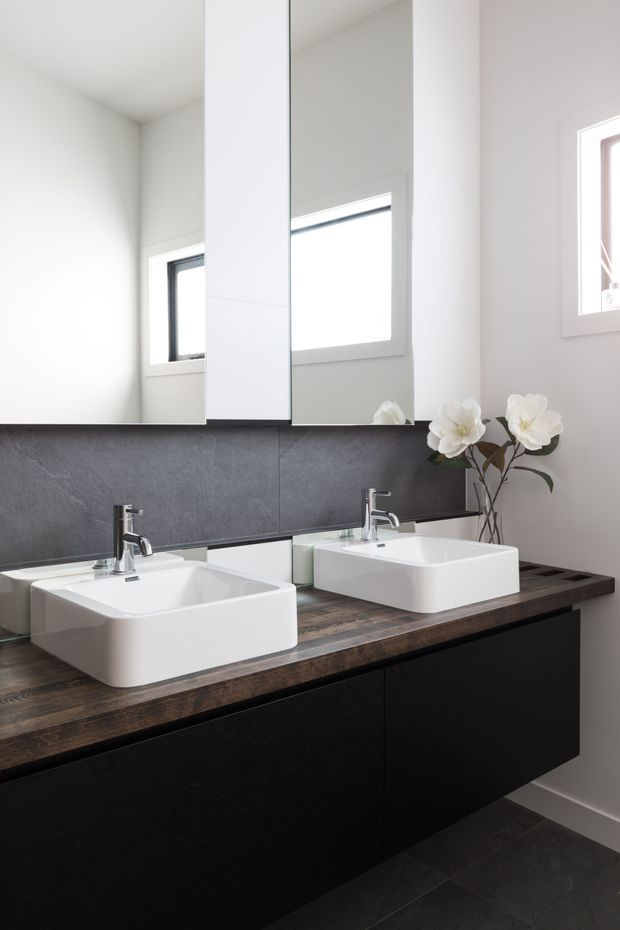 Bright contemporary bathroom vanity with two white vessel basins, chrome wall-mounted taps, dark cabinetry, a dark timber-look benchtop, and a charcoal textured wall splashback. Large mirrored panels reflect the windows, while white orchid stems add a small decorative accent. Dark floor tiles and white walls complete the clean, high-contrast scheme.