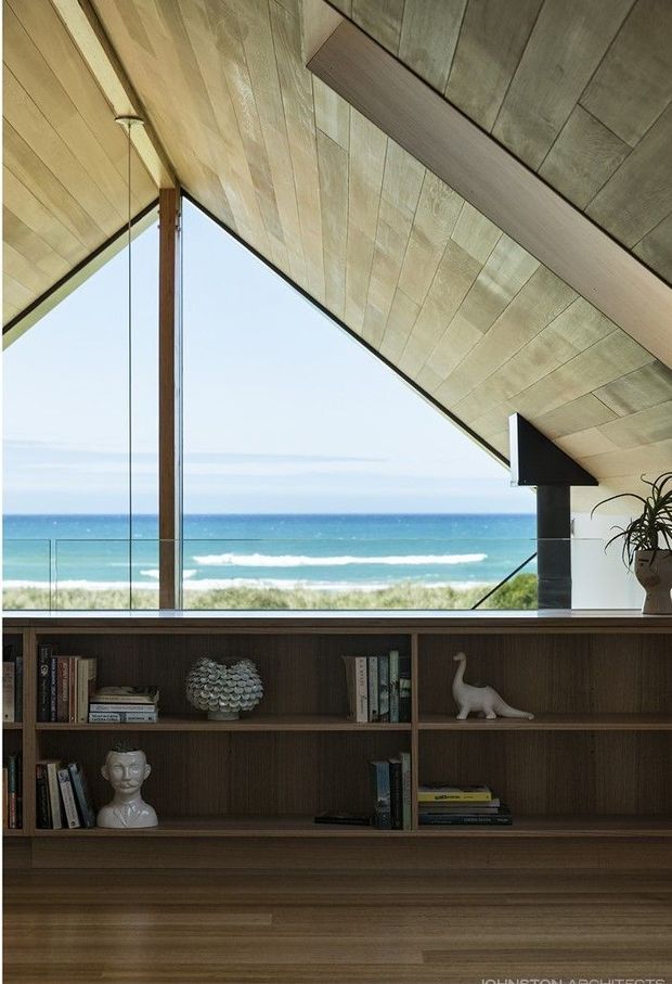 Interior view of a refined coastal living space with a dramatic sloping timber-lined ceiling, expansive floor-to-ceiling glazing, and uninterrupted ocean views. A dark timber built-in bookshelf and cabinetry spans the foreground, displaying books and sculptural objects. The room has a warm, natural material palette and strong indoor-outdoor connection.