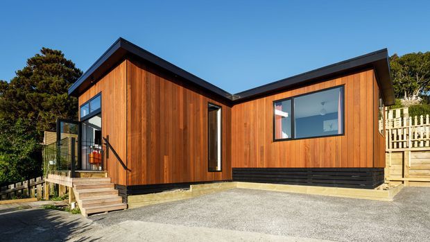 Exterior view of a compact contemporary timber-clad house with a low-pitched asymmetrical roof, black trims and base detailing, large windows, and a raised entry deck reached by steps. The home sits on a sloping, wooded suburban site with a gravel driveway and timber fencing.