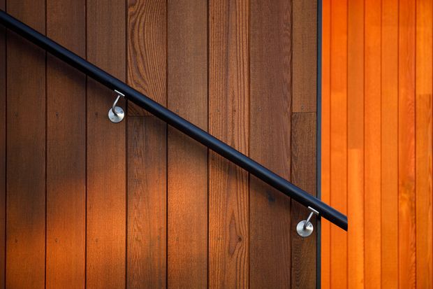 Close-up architectural detail of a black metal handrail mounted diagonally across vertically grooved dark timber wall panelling. Small silver mounting brackets and warm concealed lighting accentuate the rich wood grain and create a dramatic, warm atmosphere.