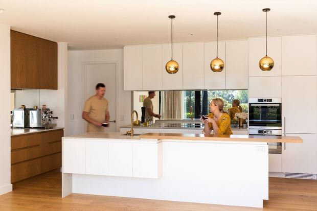 Bright contemporary kitchen with extensive white handleless cabinetry, a large central island, warm timber flooring and timber-toned cabinetry along the left wall. Three spherical brass pendant lights hang above the island, with integrated stainless-steel appliances and a mirrored splashback reflecting the room and occupants.