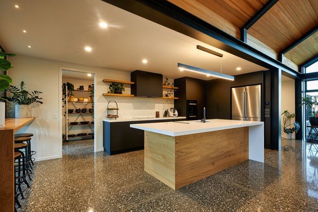 A spacious contemporary kitchen with a large light-wood island, dark cabinetry, integrated stainless-steel appliances, open shelving, and a polished aggregate or terrazzo-style floor. The kitchen is set within a high-ceilinged open-plan space with exposed timber ceiling lining, black structural beams, expansive glazing, and warm architectural lighting.
