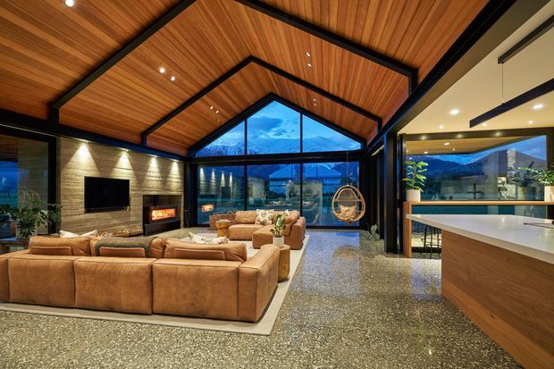 Spacious open-plan living room with a dramatic pitched timber-lined ceiling, exposed dark structural beams, floor-to-ceiling glazing, and a stone fireplace with a television. A large tan sectional seating arrangement faces the fireplace, while a contemporary kitchen with a broad island is visible on the right. The glazing reveals a mountainous landscape beyond.