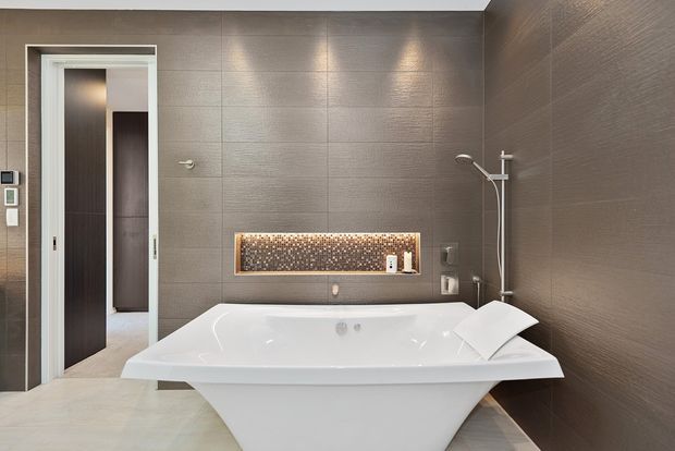 Contemporary bathroom featuring a freestanding white bathtub, wall-mounted shower, and large-format textured taupe-grey wall panels. A narrow illuminated mosaic feature niche adds warmth above the bath, with a sliding doorway opening to an adjacent space.