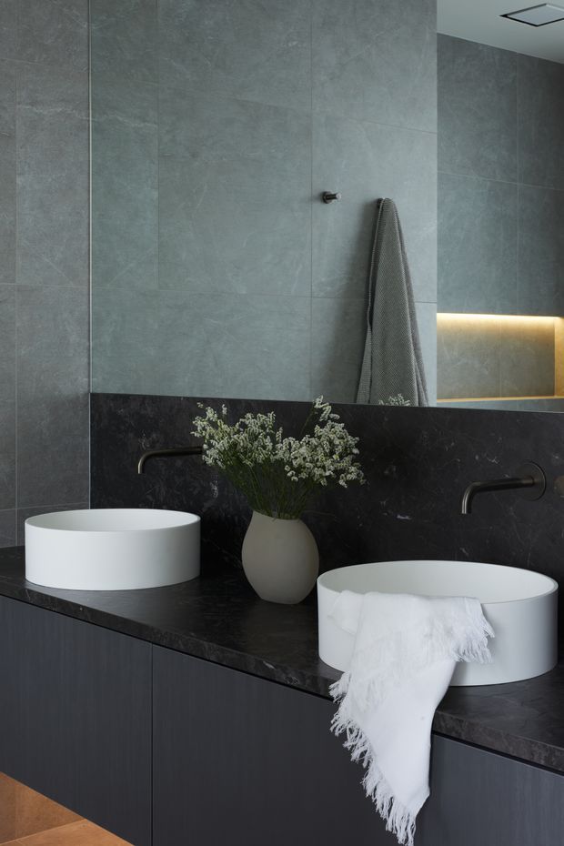 Contemporary double-vanity bathroom with two white round vessel basins on a dark stone benchtop, wall-mounted black taps, dark cabinetry, large grey stone-look wall tiles, and a wide mirror with warm concealed lighting. A folded white towel and small vase of foliage add softness to the otherwise refined, monochromatic scheme.
