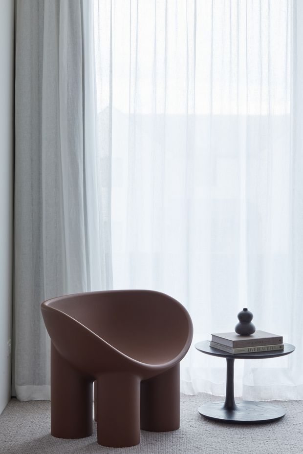 A minimalist interior vignette featuring a sculptural brown rounded armchair with cylindrical legs beside a small dark pedestal side table. The table holds stacked books and a small dark decorative vessel. Sheer white curtains diffuse abundant daylight, with grey carpet and a neutral, softly layered backdrop.