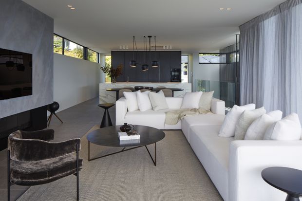 A refined contemporary open-plan living room with a large white sectional sofa, sculptural dark coffee table, armchair, and neutral textured rug. The space includes a dark fireplace and television wall, a kitchen island with pendant lighting beyond, floor-to-ceiling sheer curtains, and expansive glazing. The palette is predominantly white, grey, taupe, and black with a calm, polished atmosphere.