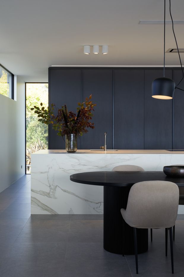 A refined contemporary kitchen and dining area with a large white veined stone island, dark charcoal full-height cabinetry, and a black oval dining table with upholstered chairs. Large windows bring in leafy garden views, while sculptural pendant lighting and integrated ceiling lights add a polished modern character.