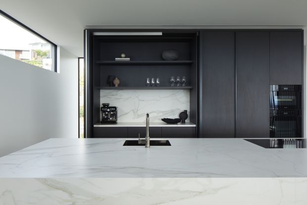 Contemporary kitchen featuring a large white marble-look island with an integrated sink and brushed metal tap. A dark charcoal built-in wall of cabinetry includes open display shelving, a marble splashback, concealed appliances, glassware, and a coffee machine. Slim black-framed glazing brings in natural light, creating a refined, high-contrast interior.