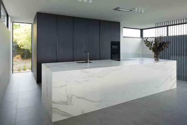 Contemporary kitchen dominated by a large white veined stone island and waterfall benchtop. The island includes an undermount sink and slim black tap, with tall dark built-in cabinetry and integrated appliances behind it. Large windows provide natural light, while a black vertical slatted screen separates the kitchen from an adjacent area. Grey tiled flooring and restrained finishes create a refined, minimalist look.